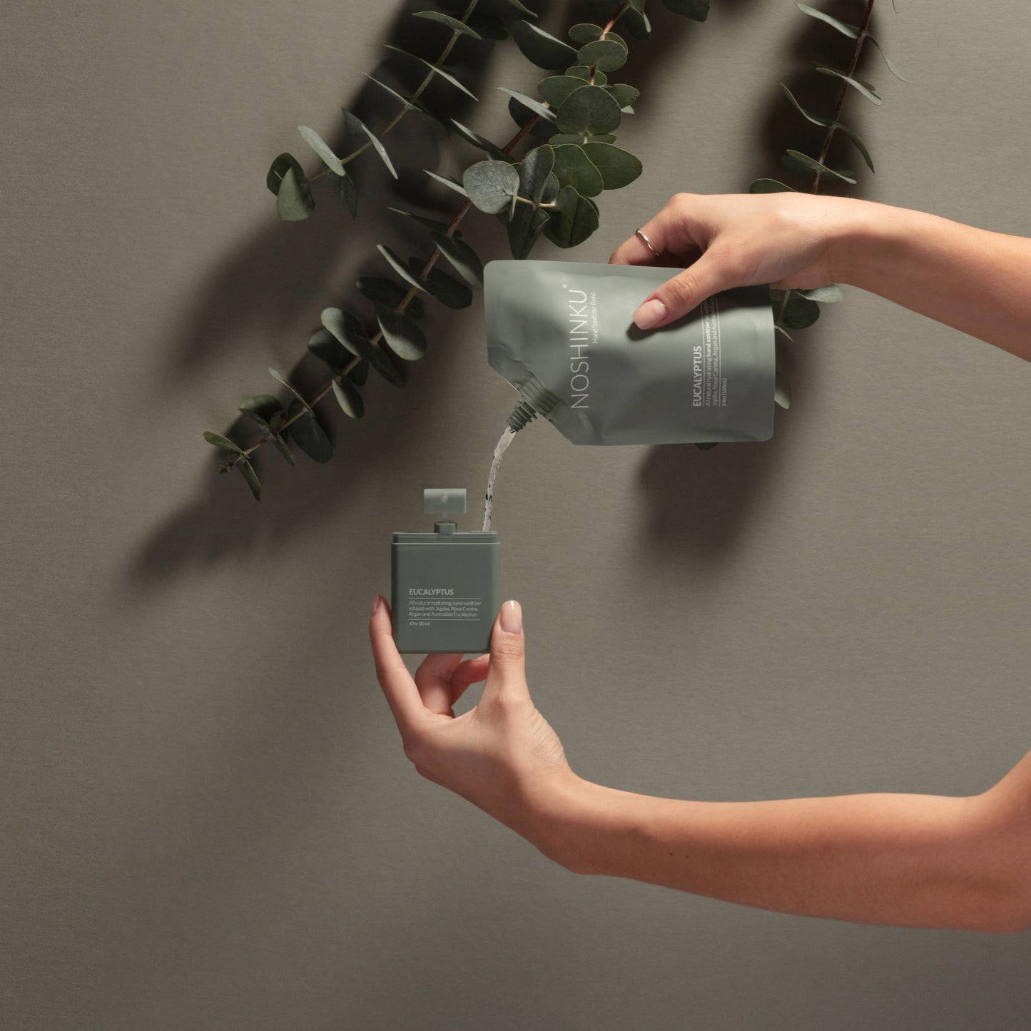 Refilling Noshinku Eucalyptus moisturizing hand sanitizer spray using a refill pouch with eucalyptus leaves in the background. Exclusively at Area Boutique.