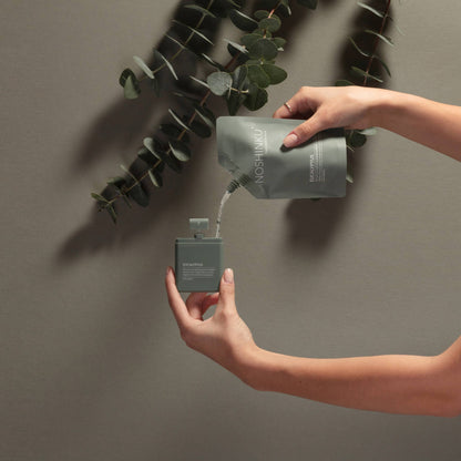 Refilling Noshinku Eucalyptus moisturizing hand sanitizer spray using a refill pouch with eucalyptus leaves in the background. Exclusively at Area Boutique.