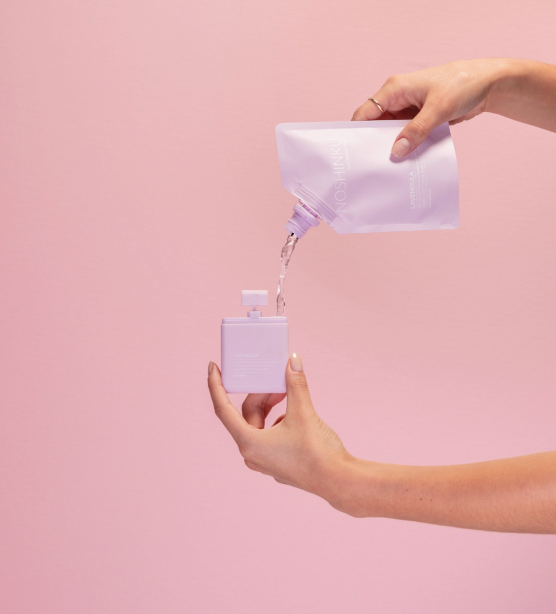 Refilling Noshinku Lavendula moisturizing hand sanitizer spray using a refill pouch against a soft pink background. Exclusively at Area Boutique.