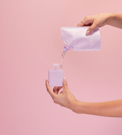 Refilling Noshinku Lavendula moisturizing hand sanitizer spray using a refill pouch against a soft pink background. Exclusively at Area Boutique.