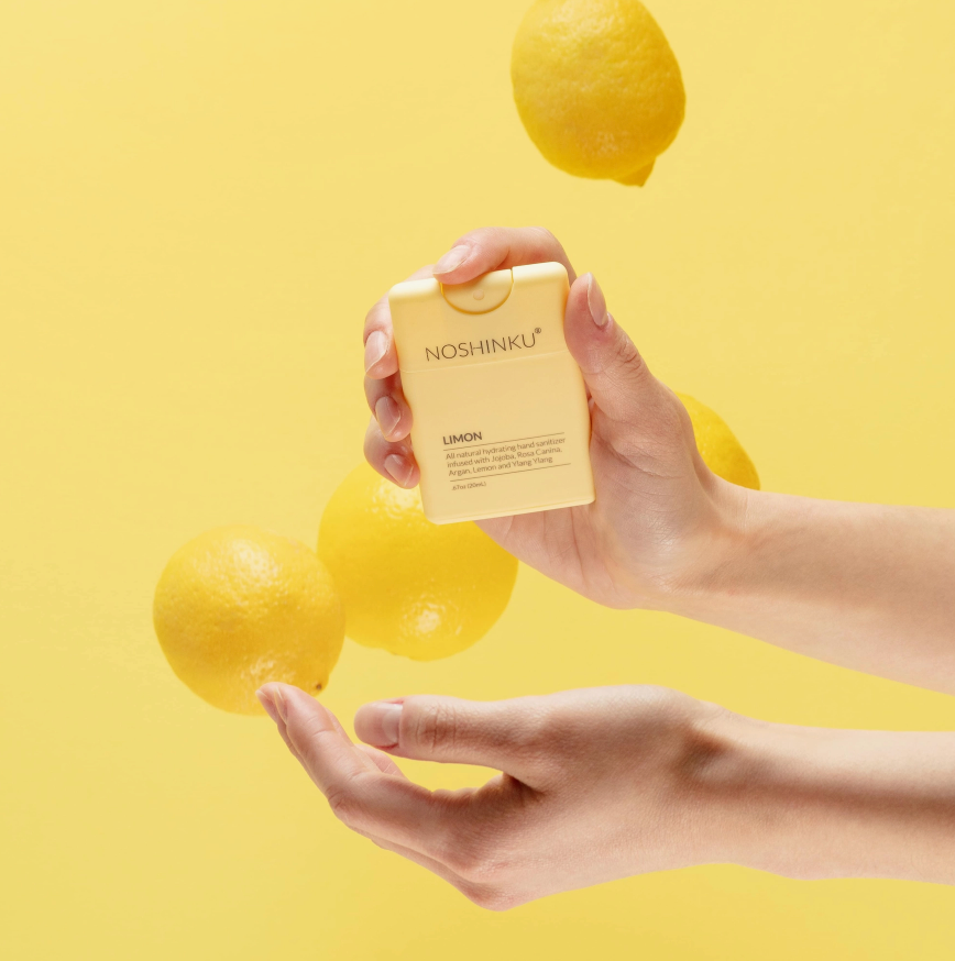 Hands holding Noshinku Lemon refillable pocket hand sanitizer surrounded by fresh lemons against a bright yellow background highlighting its uplifting citrus scent. Exclusively at Area Boutique.