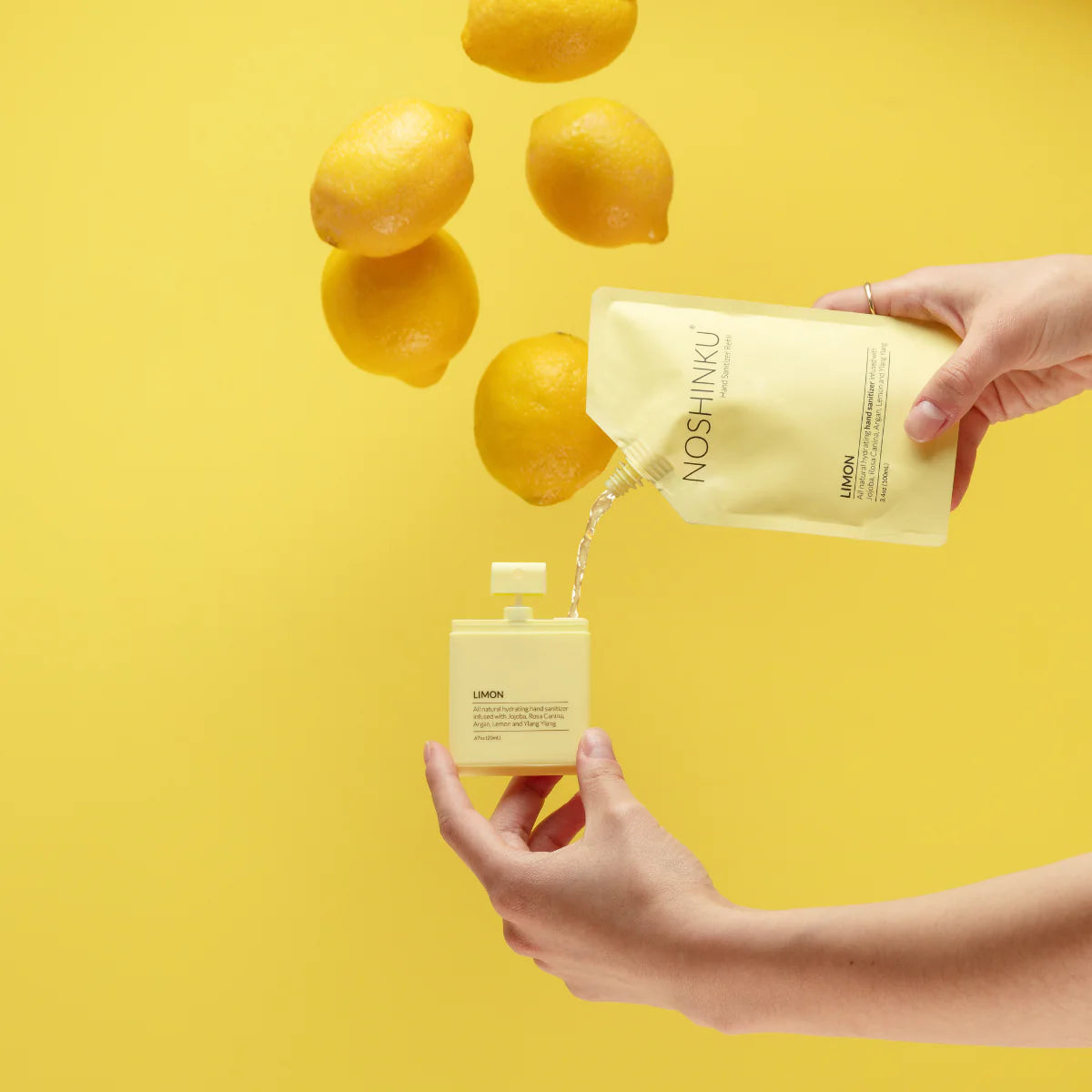 Noshinku Lemon refill pouch pouring hydrating hand sanitizer into a refillable pocket spray bottle surrounded by fresh lemons against a bright yellow background. Exclusively at Area Boutique.