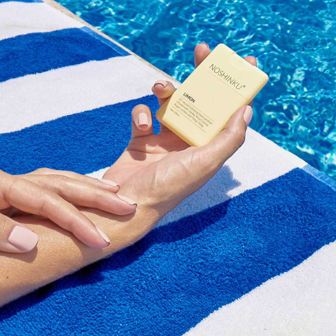 Noshinku Lemon refillable pocket hand sanitizer held poolside on a striped towel featuring a bright citrus scent and travel-friendly summer-ready design. Exclusively at Area Boutique.