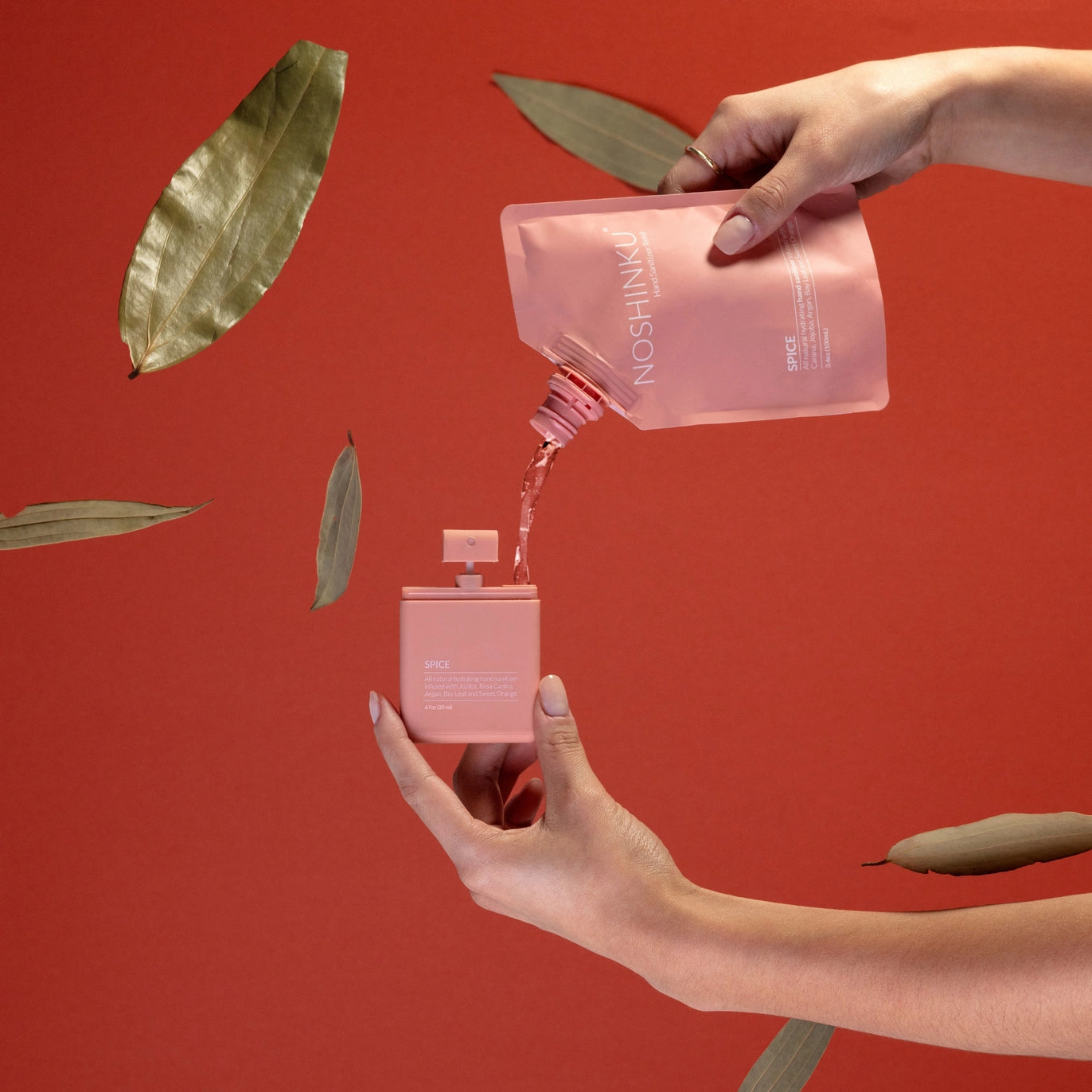 Refilling Noshinku Spice moisturizing hand sanitizer spray using a refill pouch with bay leaves against a warm red background. Exclusively at Area Boutique.