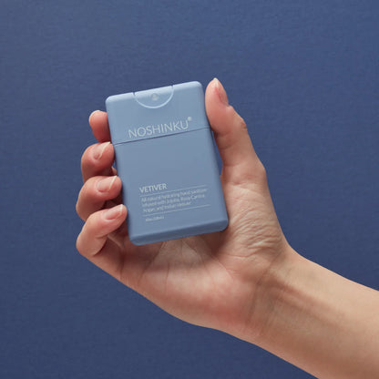Hand holding the Noshinku Vetiver refillable pocket hand sanitizer spray in a matte blue slim bottle against a deep blue background.