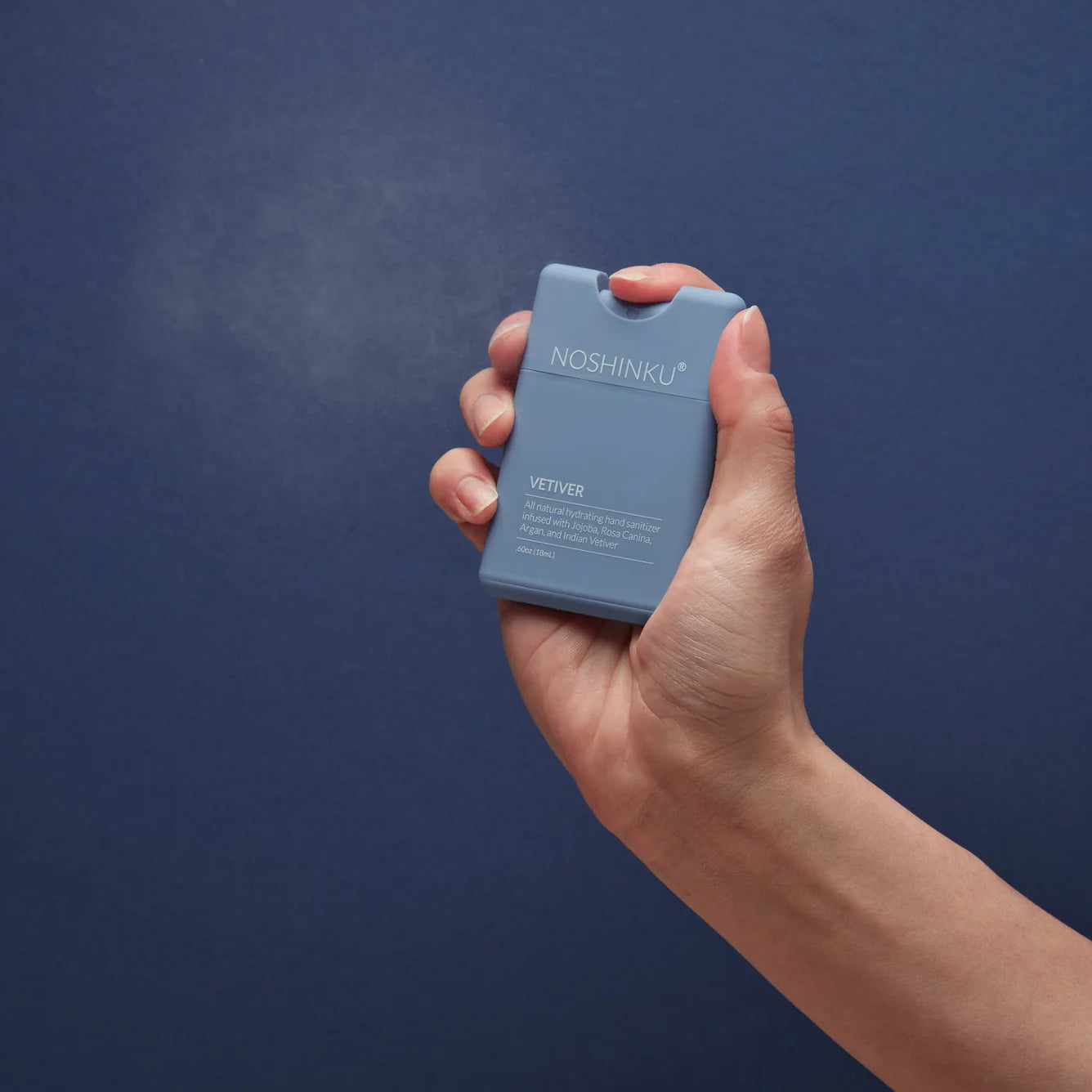 Person spraying Noshinku Refillable Hand Sanitizer Spray – Vetiver from a slim matte blue pocket sanitizer bottle against a blue background.