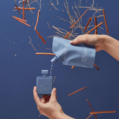 Refilling Noshinku Refillable Hand Sanitizer Spray – Vetiver using a refill pouch with woody vetiver-inspired elements against a deep blue background.